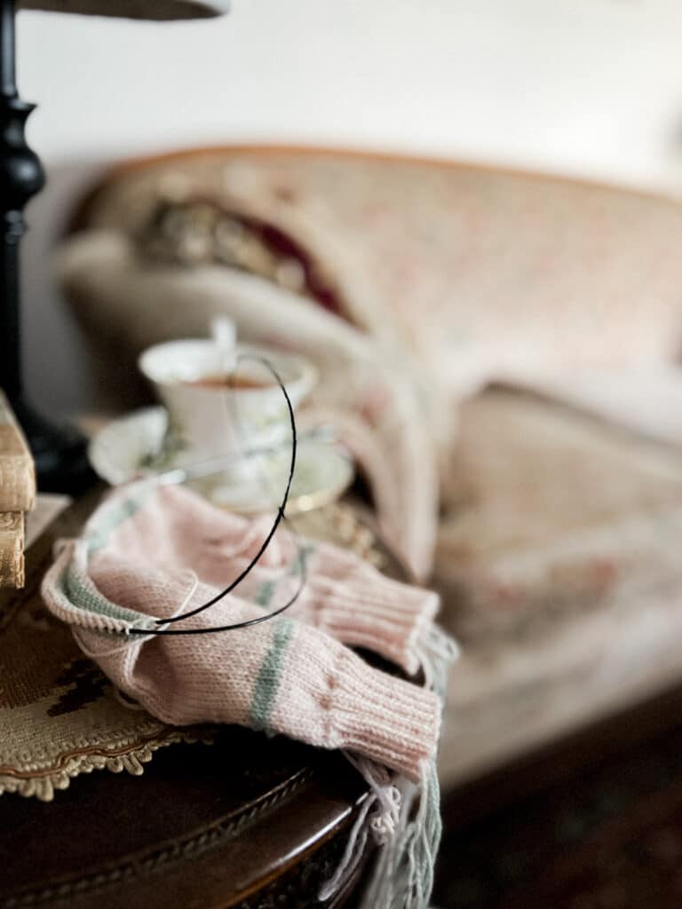 A pair of pale pink mittens with light green stripes in progress on a long set of circular needles sits on a small side table next to a sofa (blurred in the background).
