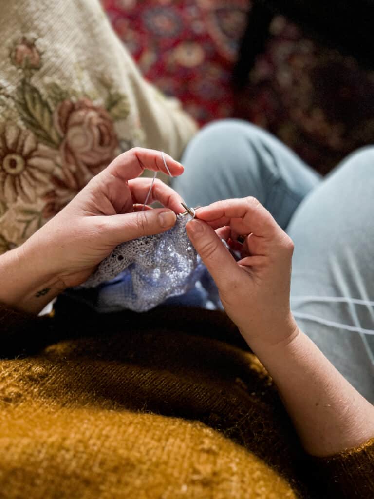 A white woman wearing a deep mustard-colored sweater works on knitting a light blue shawl. The photo is taken from overhead, showing her hands in the position she'd see them as she knits.