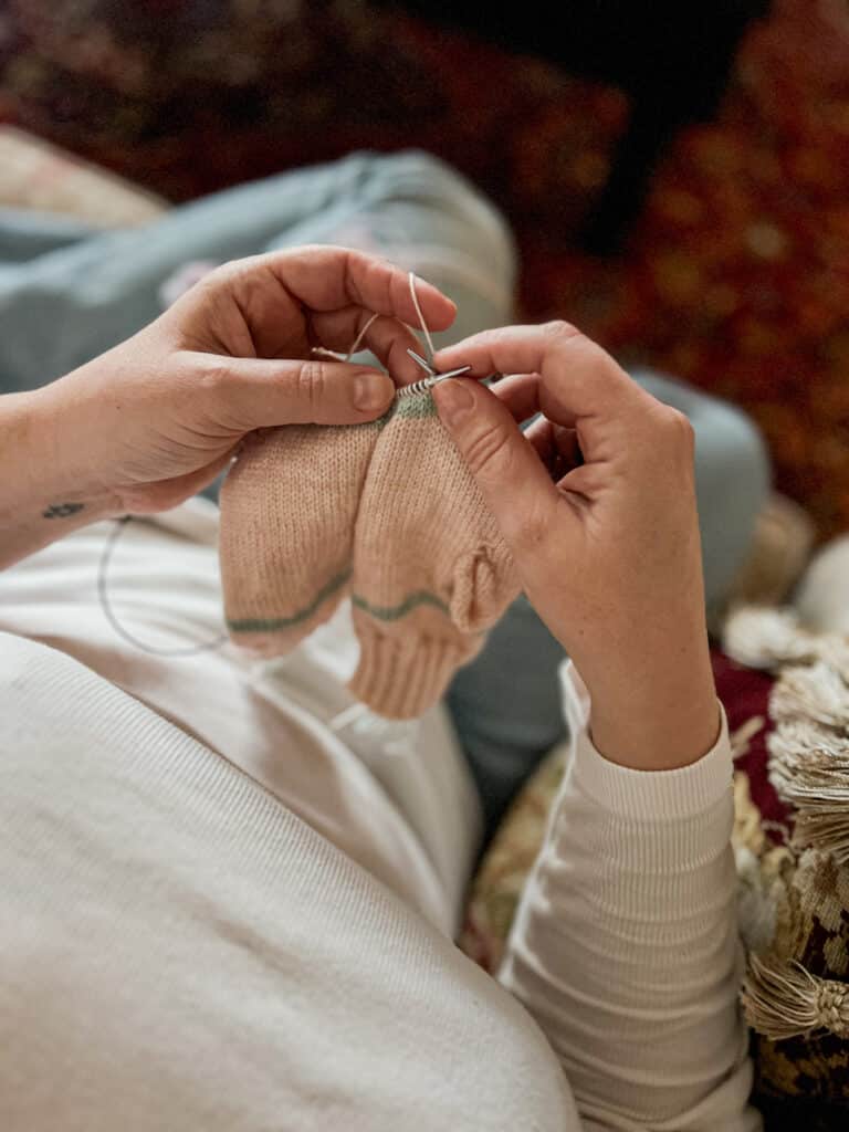 A white woman wearing a white t-shirt works on knitting a pair of pale pink mittens with light green stripes. The photo is taken from overhead, showing her hands in the position she'd see them as she knits.
