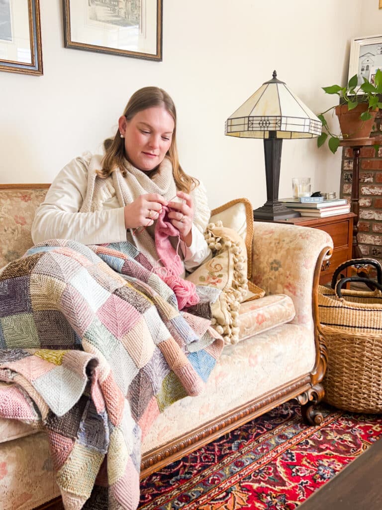 A white woman with shoulder-length blonde hair knits a pink sock while snuggled up under a multicolored knit blanket made up of mitered squares. She is lounging with her feet tucked under her on a vintage sofa with faded floral upholstery.