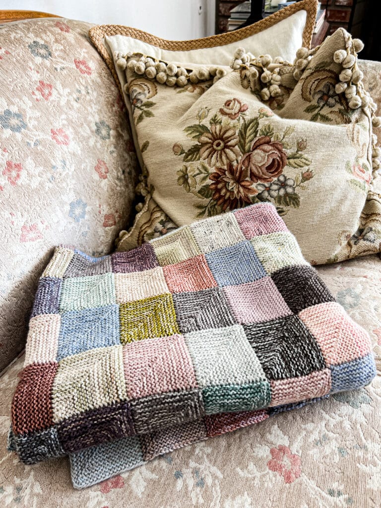 A folded knit blanket made up of multicolored mitered squares sits on a vintage sofa with faded floral upholstery.