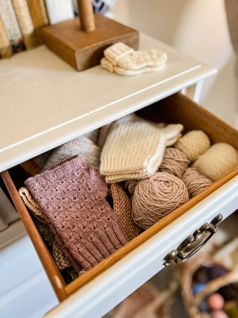 An open drawer of a white dresser reveals two pastel pairs of handknit mitts and several small balls of pastel yarn leftovers.