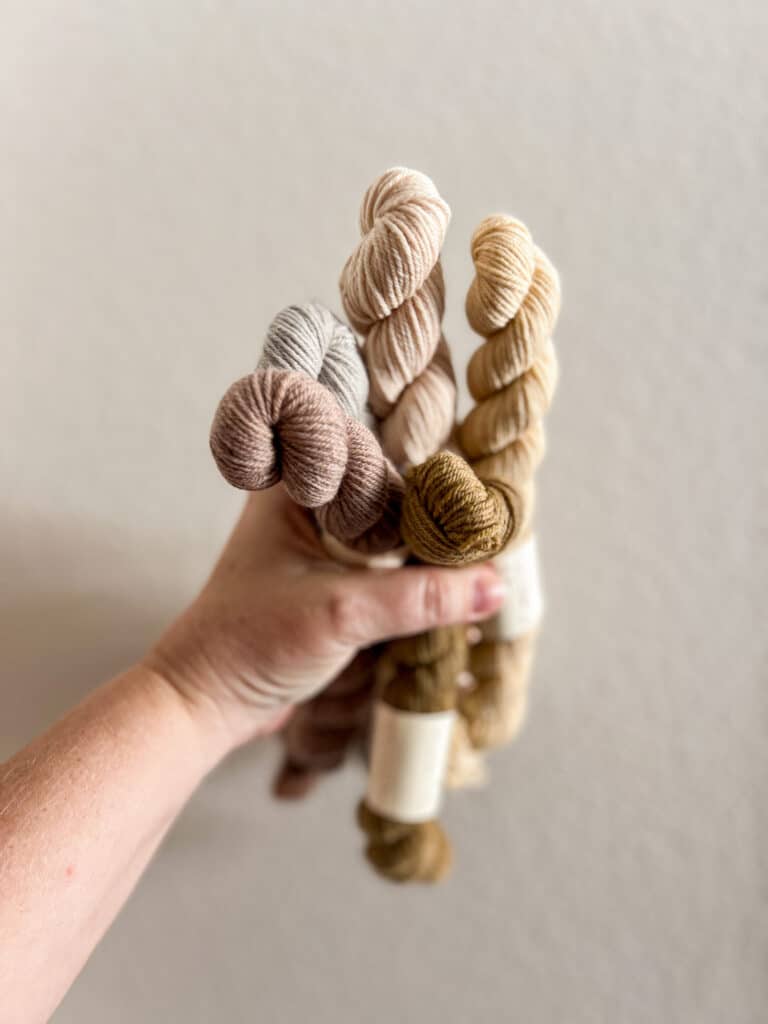 A white woman's left hand grips a bundle of pastel mini skeins of yarn in front of a white wall.