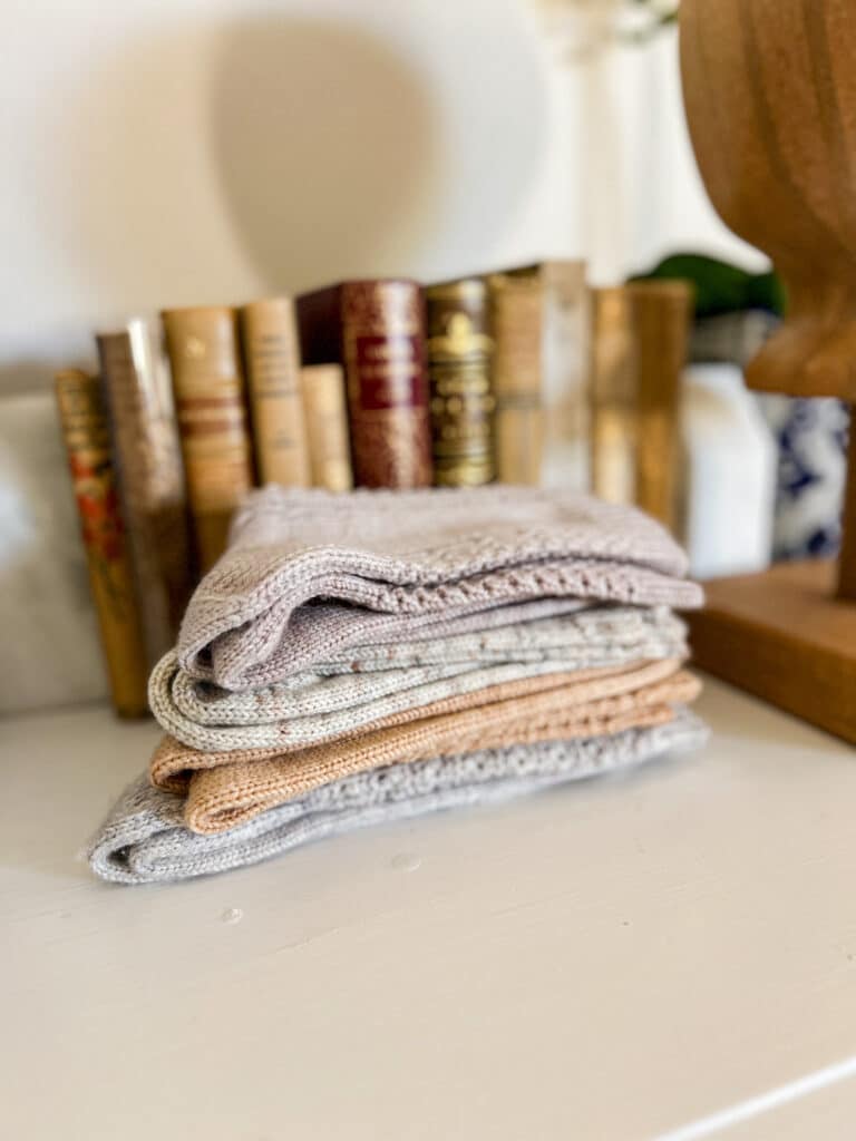Four pairs of pastel, handknit socks are folded and stacked on top of a white dresser in front of some antique books.