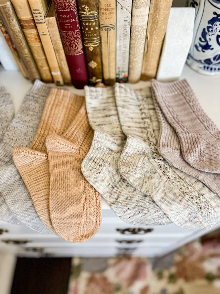 Four pairs of pastel, handknit socks are laid out across the top of a white dresser with some antique books in the background.