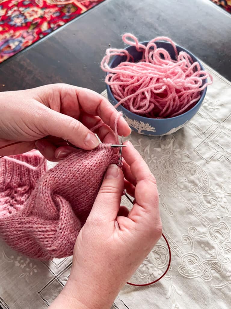 A white woman's hands work on a pink worsted-weight sock using the Magic Loop method.