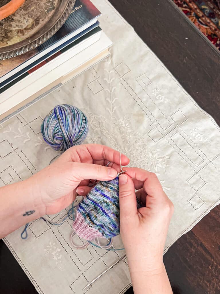 A white woman's hands knit a blue and purple multicolored sock on a pair of steel knitting needles.