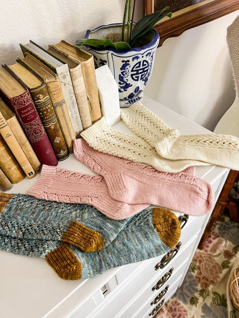 Three pairs of handknit socks in shades of blue, pink, and white sit on top of a white dresser with antique books in the background. The photo is taken from a low angle near the heels of the closest pair of socks.