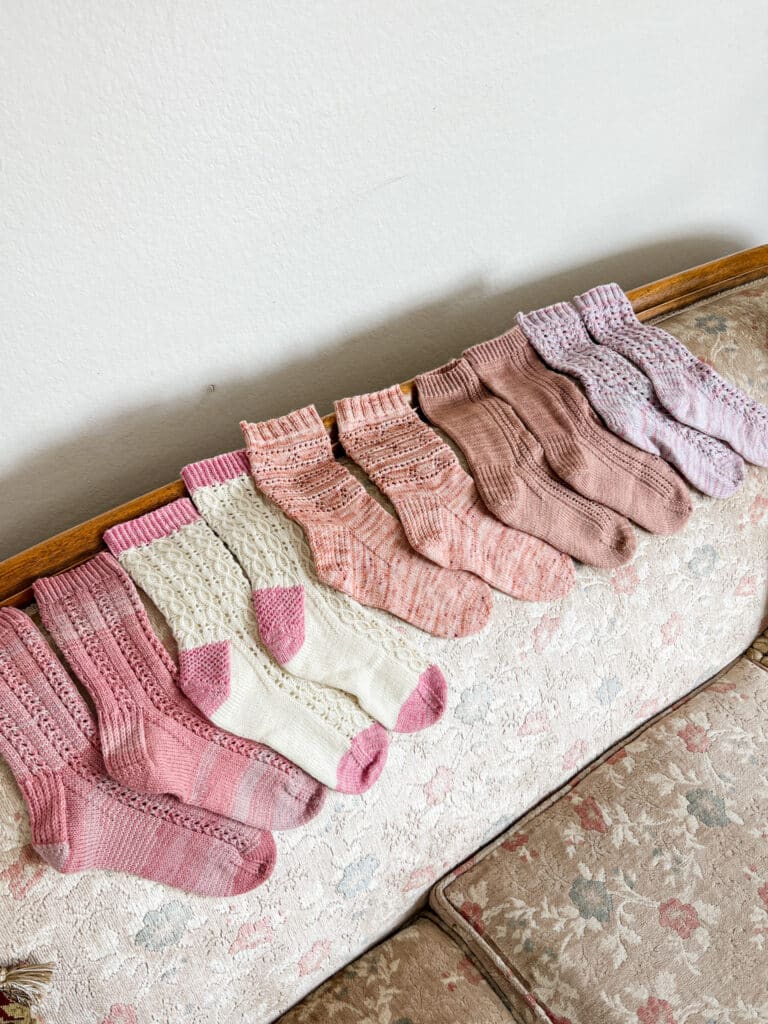 Five pairs of handknit socks in various shades of pink and white are laid out across the back of a vintage sofa with floral upholstery.