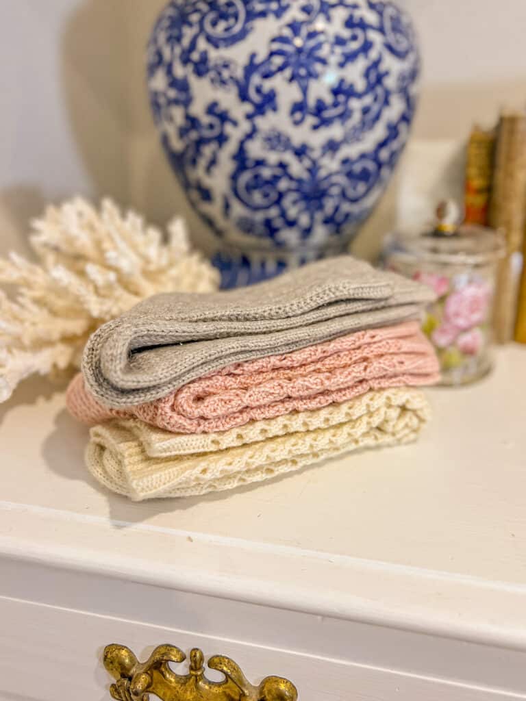 Three pairs of folded, handknit socks sit on top of a white dresser in front of a blue and white lamp and a large chunk of white coral.