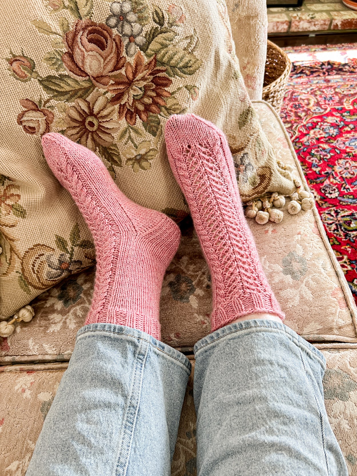 A pair of feet wearing thick pink socks extend from the bottoms of a pair of light blue jeans. The socks have front panels of lace and cables.