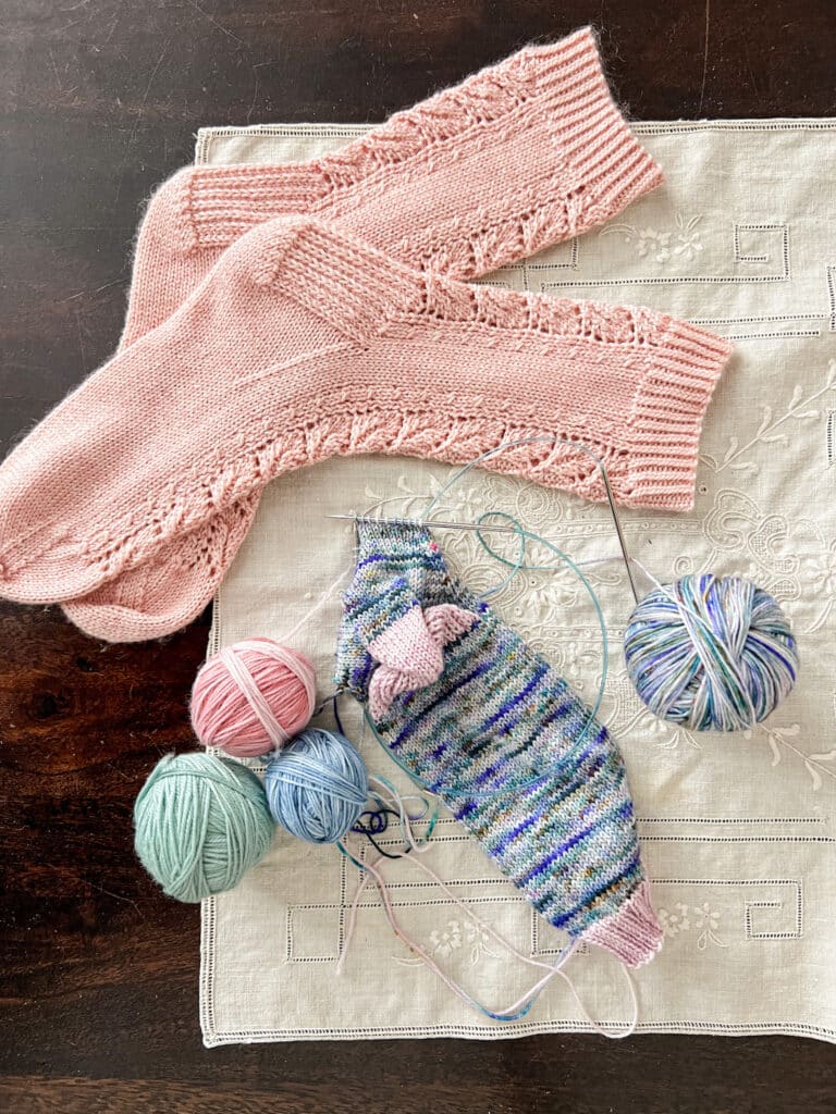 A blue multicolored sock in progress on two circular needles is laid out on a table top next to some finished pink socks and a few balls of pastel yarn.