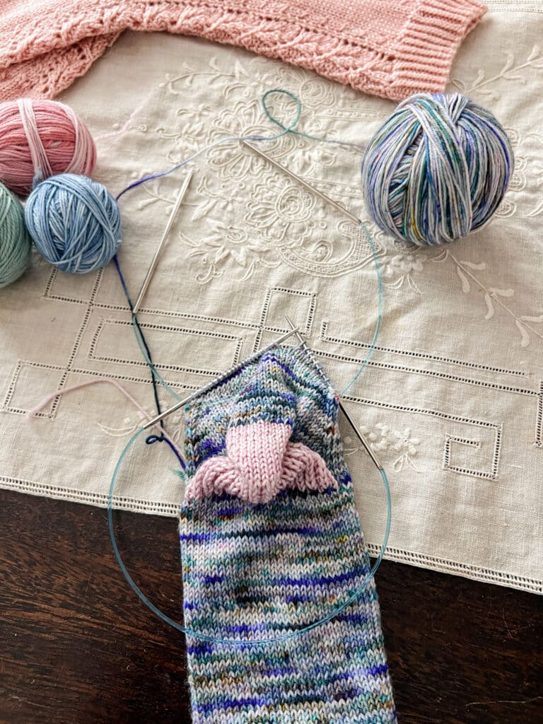 A handknit sock in progress on two circular needles is laid out on a tabletop with the needles angled upward to show how one sits on the front half of the stitches and one sits on the back.