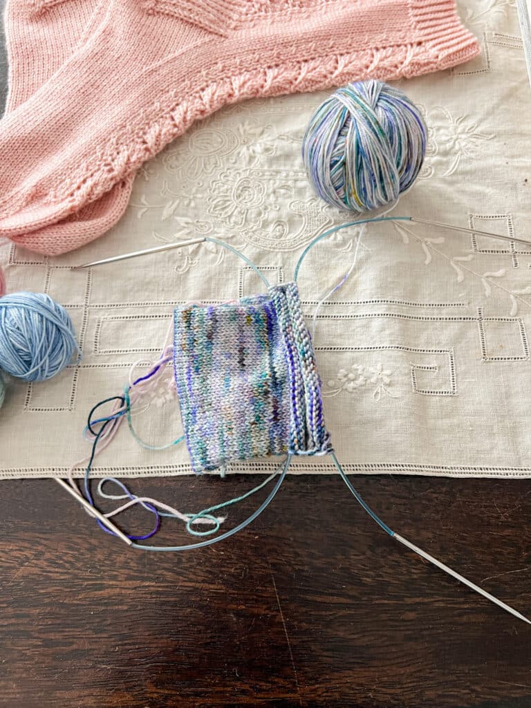 A handknit sock in progress on two circular needles is laid out sideways on a table with the end angled upward to better show how the stitches are divided into two halves across the two needles.