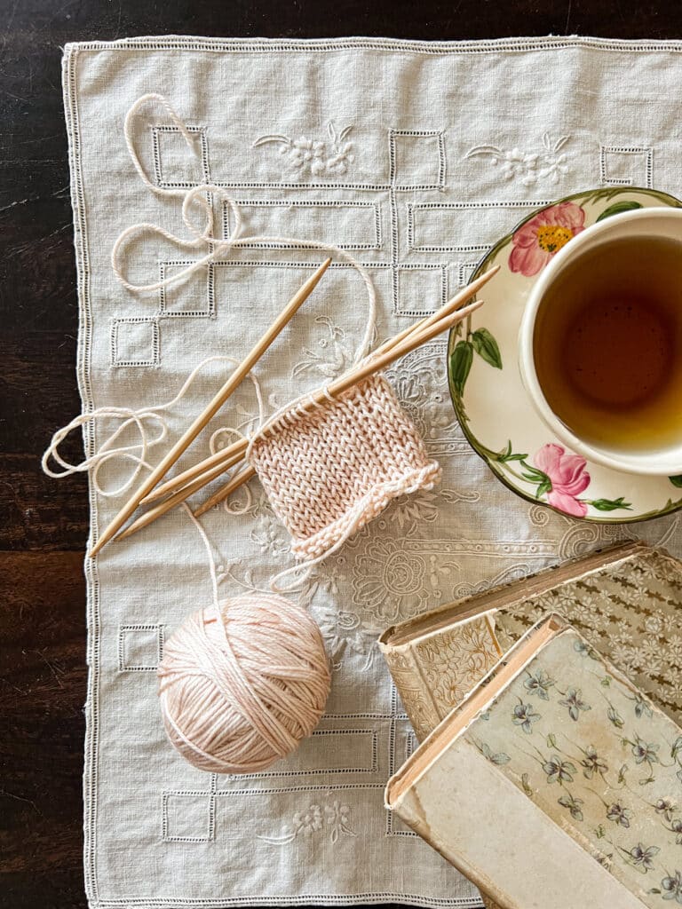 A top-down photo showing a tube of pale peach knitting on wooden DPNs. The knitting has significant laddering at the spot where the needles join. Around the knitting are a teacup filled with tea and some antique books.