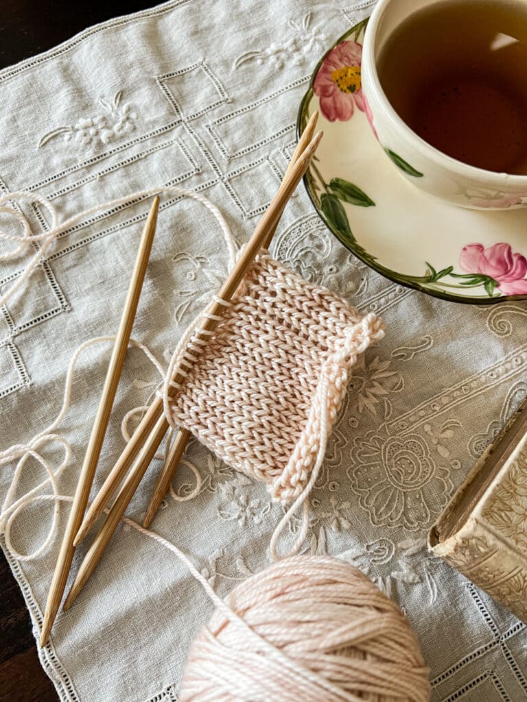 A low-angle photo showing a tube of pale peach knitting on wooden DPNs. The knitting has significant laddering at the spot where the needles join. Around the knitting are a teacup filled with tea and some antique books.