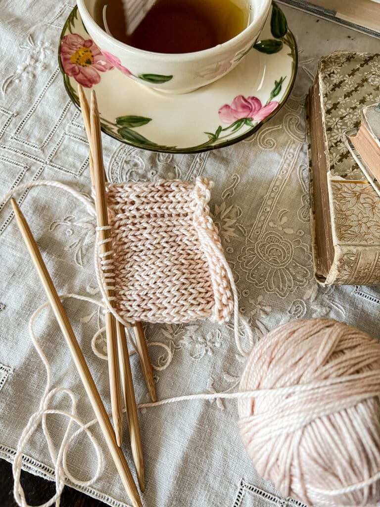 A low-angle photo zoomed in on a tube of pale peach knitting on wooden DPNs. The knitting has significant laddering at the spot where the needles join. Around the knitting are a teacup filled with tea and some antique books.