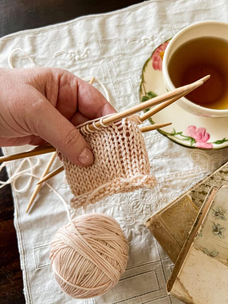 A white woman's left hand holds a tube of pale peach knitting with significant laddering issues at the spot where the needles join.