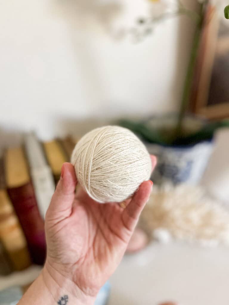A white woman's left hand holds a white ball of yarn.
