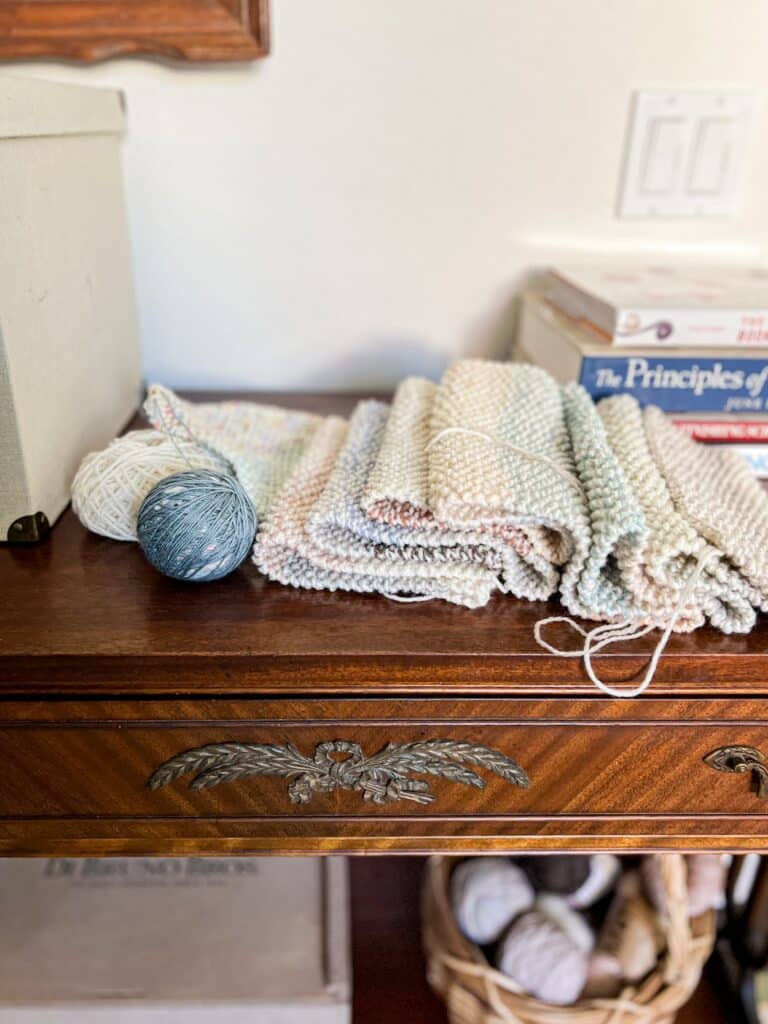 A multicolored seed stitch scarf on knitting needles, still in progress, sits on top of an antique mahogany table with a ball of scrap yarn next to it.