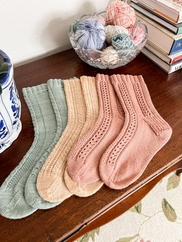 Three pairs of handknit socks in green, tan, and pink are laid out on top of a dark wooden table with the toes pointing to the left.