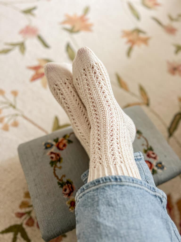 A pair of feet wearing white, lacy, handknit socks rests on top of a vintage needlepoint stool with flowers embroidered on it. The feet are crossed at the ankles.