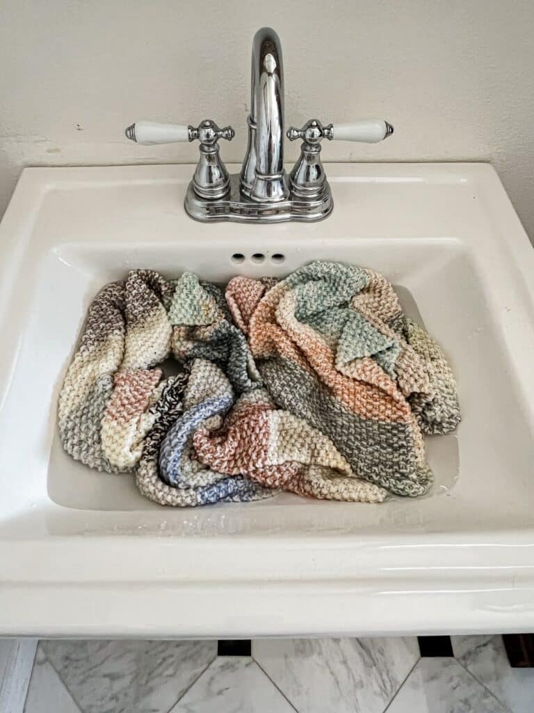 A multicolored seed stitch scarf soaks in a white sink with polished chrome and porcelain fixtures.