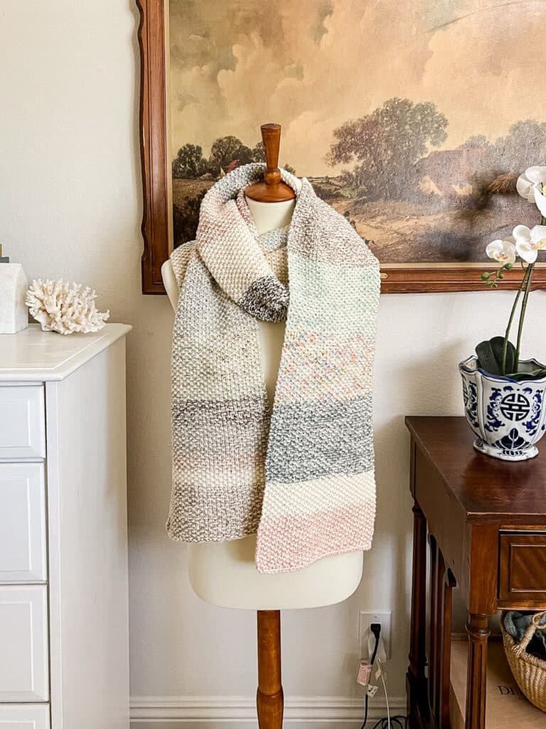 A straight-on photo of a multicolored seed stitch scarf draped around the neck of a white dressmaker's form. The form stands between a white vintage dresser and a mahogany console table. Filling most of the wall behind the dressmaker's form is a vintage landscape art print in a carved wooden frame.