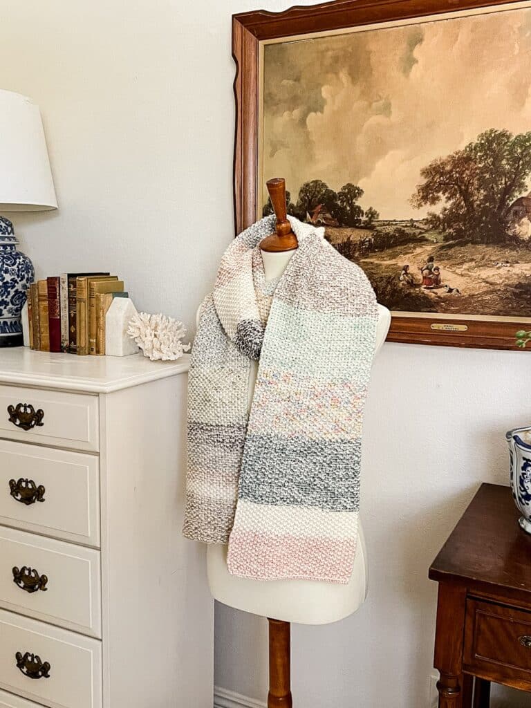 A side-angle image of a multicolored seed stitch scarf draped around the neck of a white mannequin form. In the background are a vintage art print in a carved wooden frame and a white dresser with lots of antique books on top.