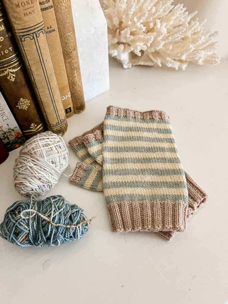 A pair of striped fingerless mitts and two small balls of yarn sit on top of a white surface with several antique books in the background.
