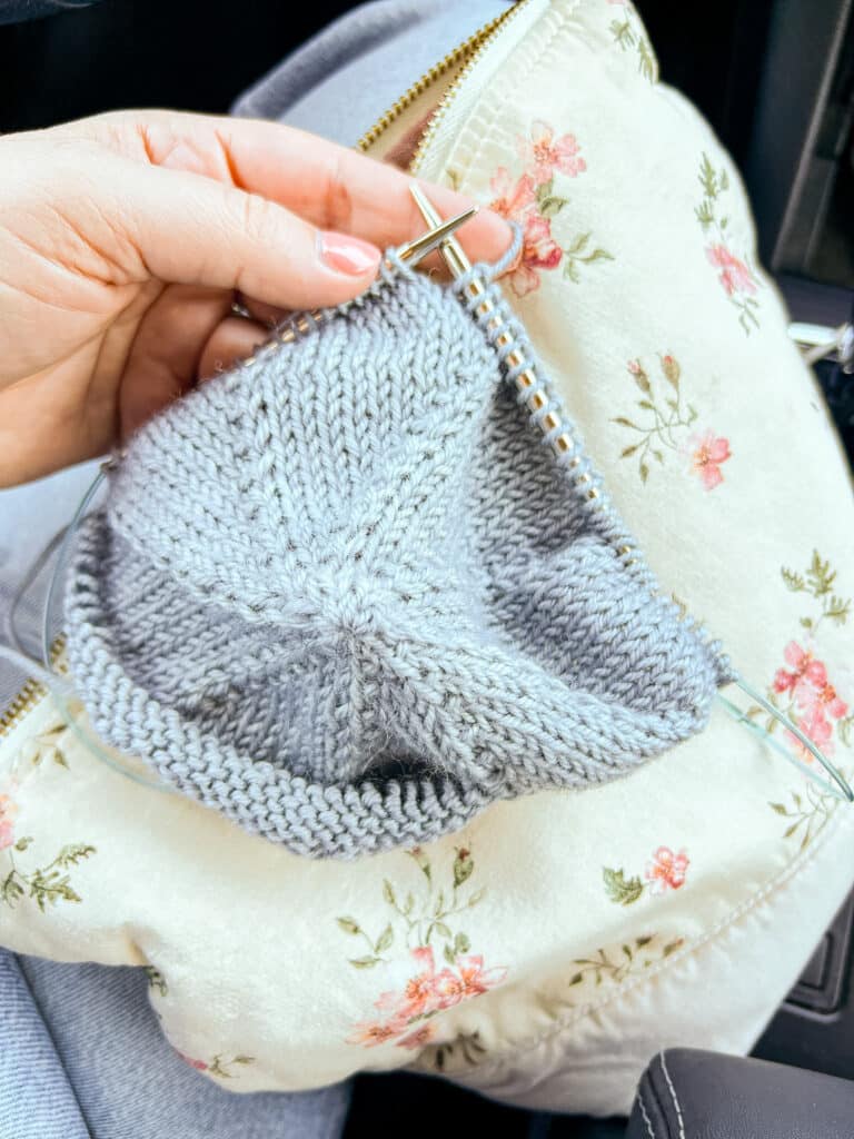 A white woman's left hand holds a light blue knit hat in progress on a set of steel circular knitting needles.