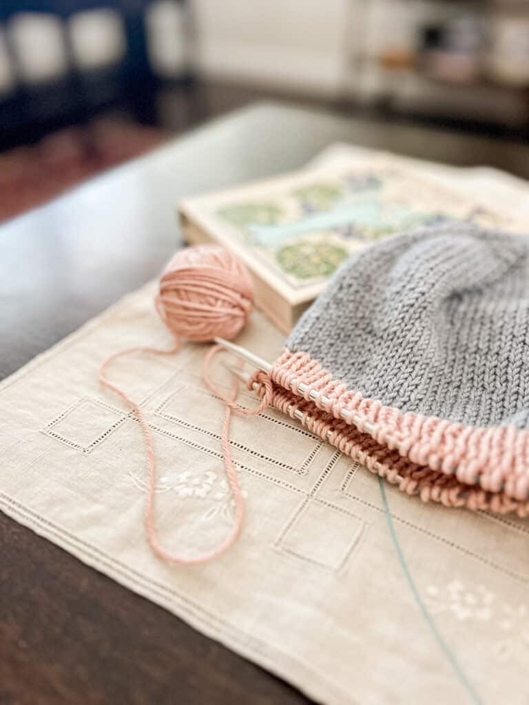 A light blue hat with a pink brim is on a set of steel knitting needles sitting on a dark wood coffee table.