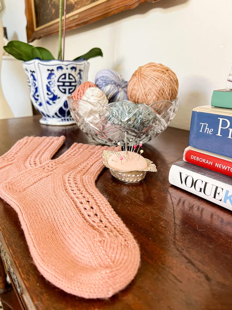 A pair of pink handknit socks is laid out on a wooden tabletop. Nearby are a bowl of leftover yarn balls and a pink pincushion filled with pins.