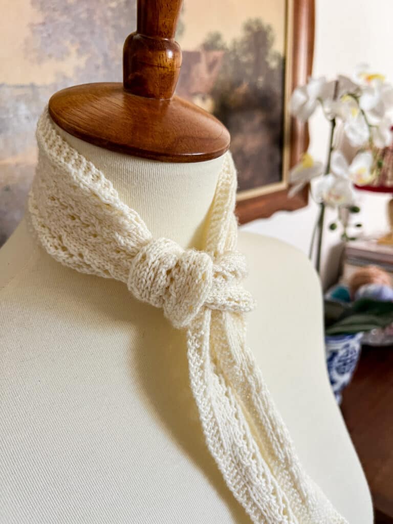 A small, white scarflet with eyelet details is tied around the neck of a white dressmaker's form.