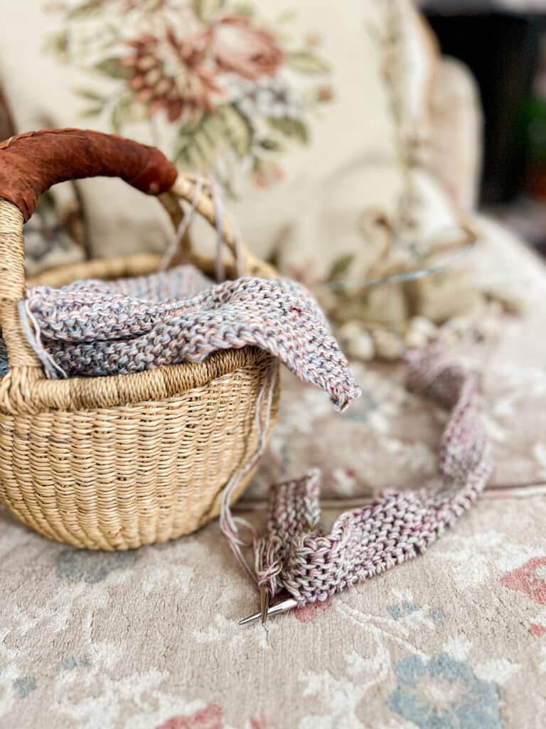 A small woven basket holds one large finished mitered square as it sits on a faded floral sofa cushion next to another mitered square in progress on a set of steel needles.