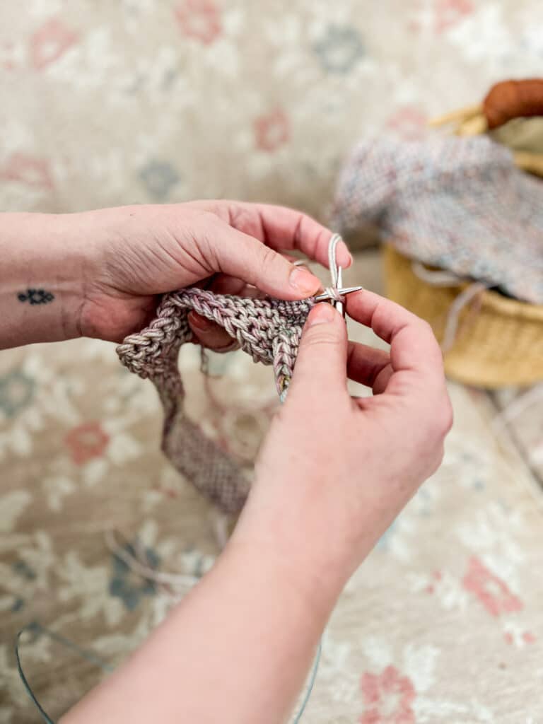 A white woman's hands hold steel knitting needles and work on knitting a light purple mitered square.