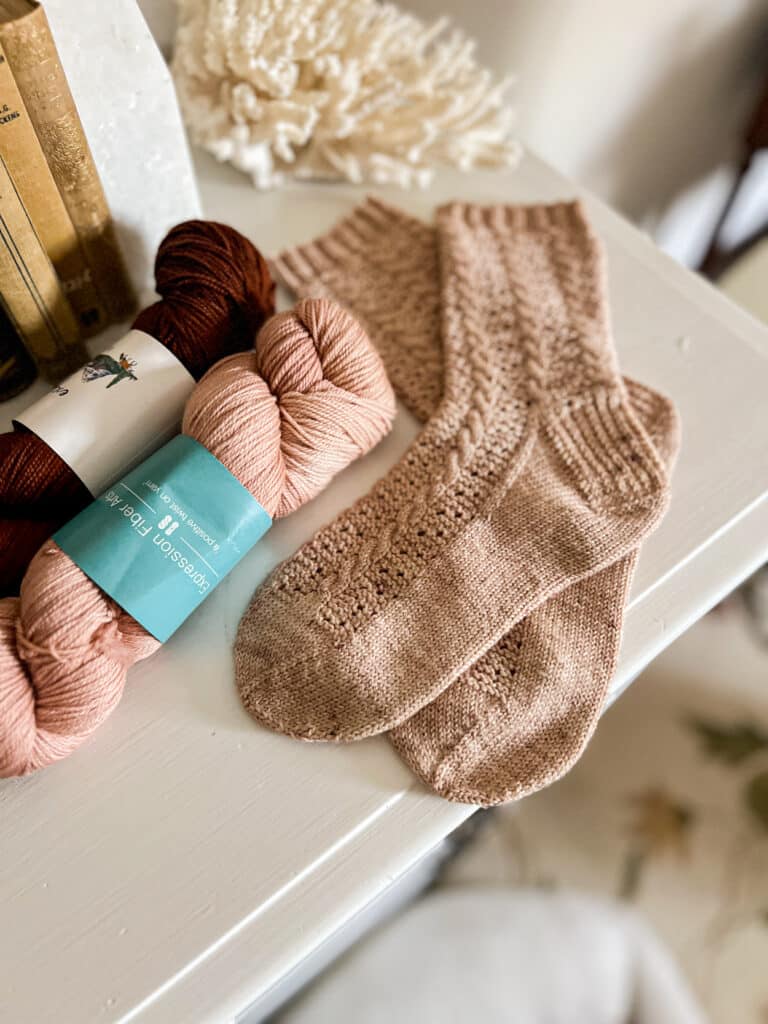 A pair of handknit cabled socks in a dusty pink color sits on top of a white dresser next to a skein of darker pink yarn and a skein of deep burgundy yarn.