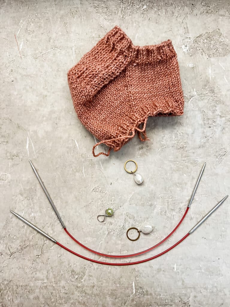 A pair of Addi Sock Wonder Needles (top) compared to a pair of ChiaoGoo Shorties (bottom). This shows how the Sock Wonder has a longer needle tip on one side compared to the ChiaoGoo Shorties. The needles are sitting next to some stitch markers and a finished knit sock heel in pink.