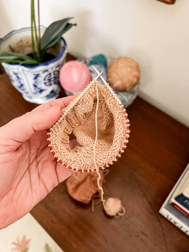 A white woman's left hand holds some small-circumference knitting on progress on its tiny circular knitting needles. The needles have a longer tip on the right hand side than on the left hand side.