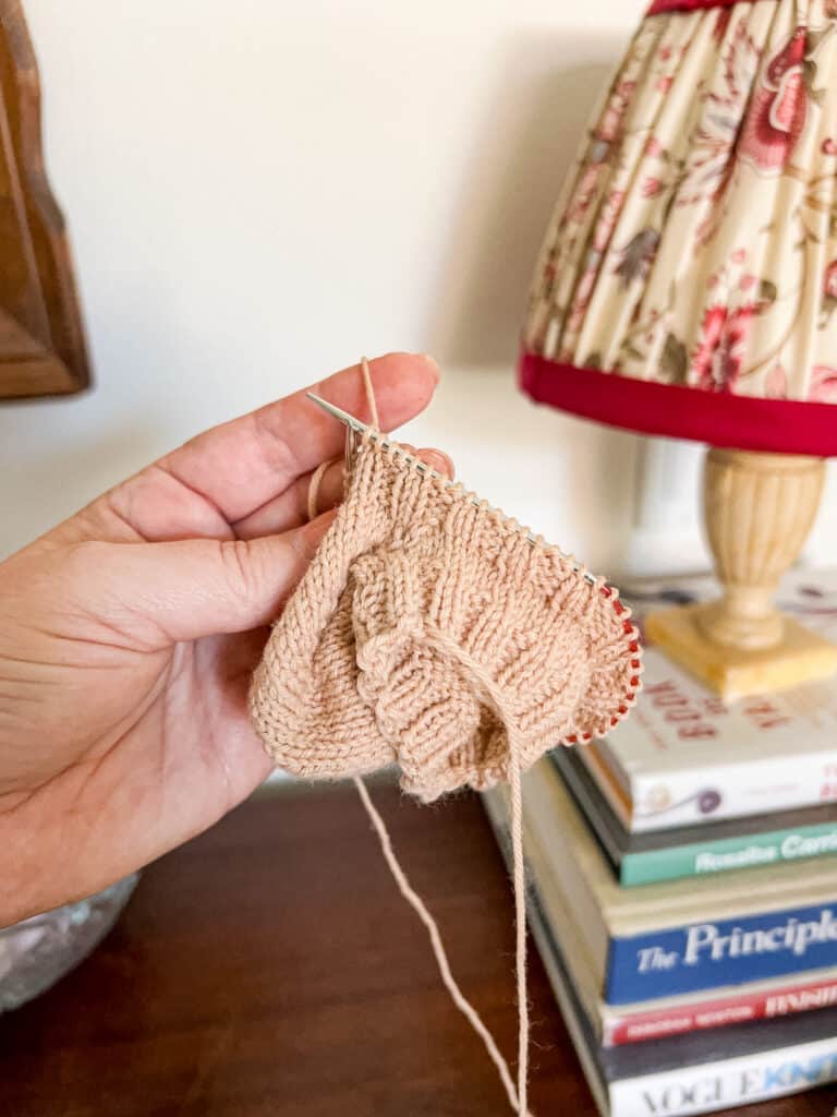 A white woman's left hand holds a small piece of knitting in progress on tiny circular needles. The work is angled so the viewer can more easily see the longer needle tip on the right hand side.
