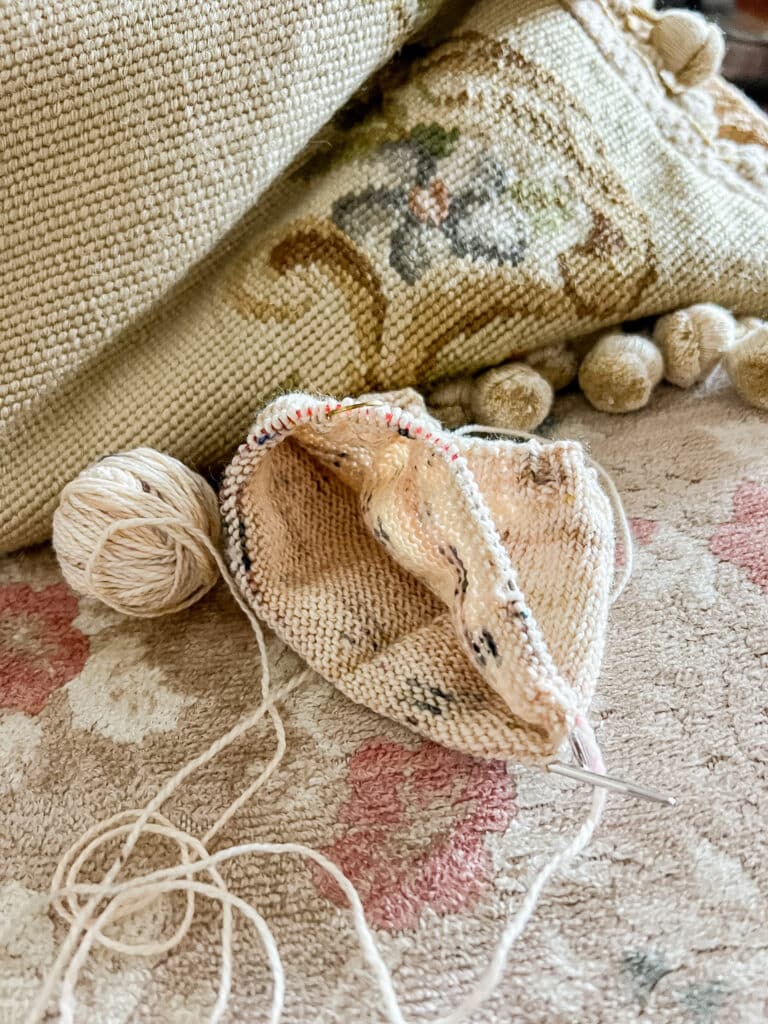 An in-progress cream sock with speckles on an Addi Sock Wonder needle sits on a vintage sofa with faded floral upholstery.