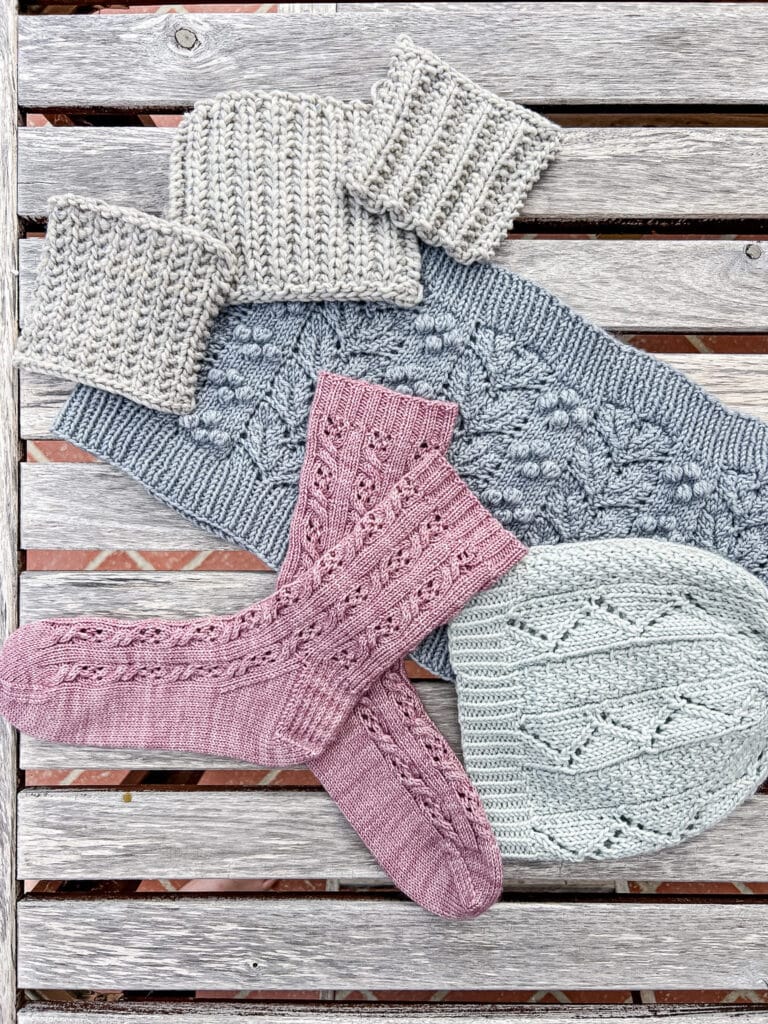 Three gray swatches of different ribbing styles sit on a weathered outdoor wooden table along with some pink socks, a blue cowl, and a gray-green hat.