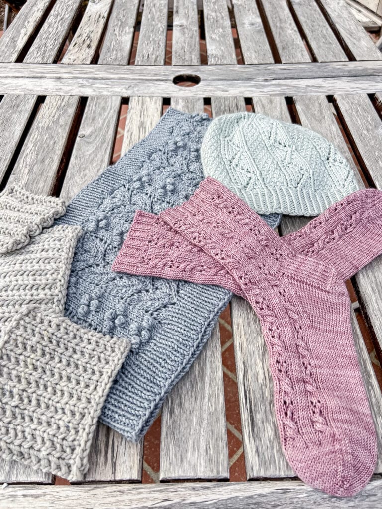 A low, side-angle photo showing three gray swatches of different ribbing styles sitting on a weathered outdoor wooden table along with some pink socks, a blue cowl, and a gray-green hat.
