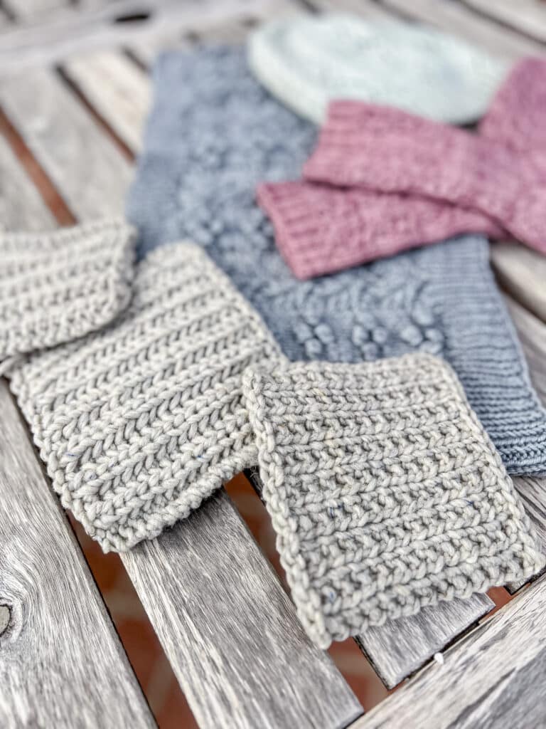 A low, side-angle photo showing three gray swatches of different kinds of ribbing on a weathered gray outdoor wooden table. Blurred in the background are additional pastel handknits.