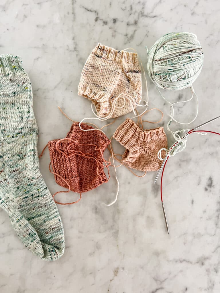 Several handknit samples of different types of sock heels, along with a finished green sock, are laid out flat on a white marble countertop.