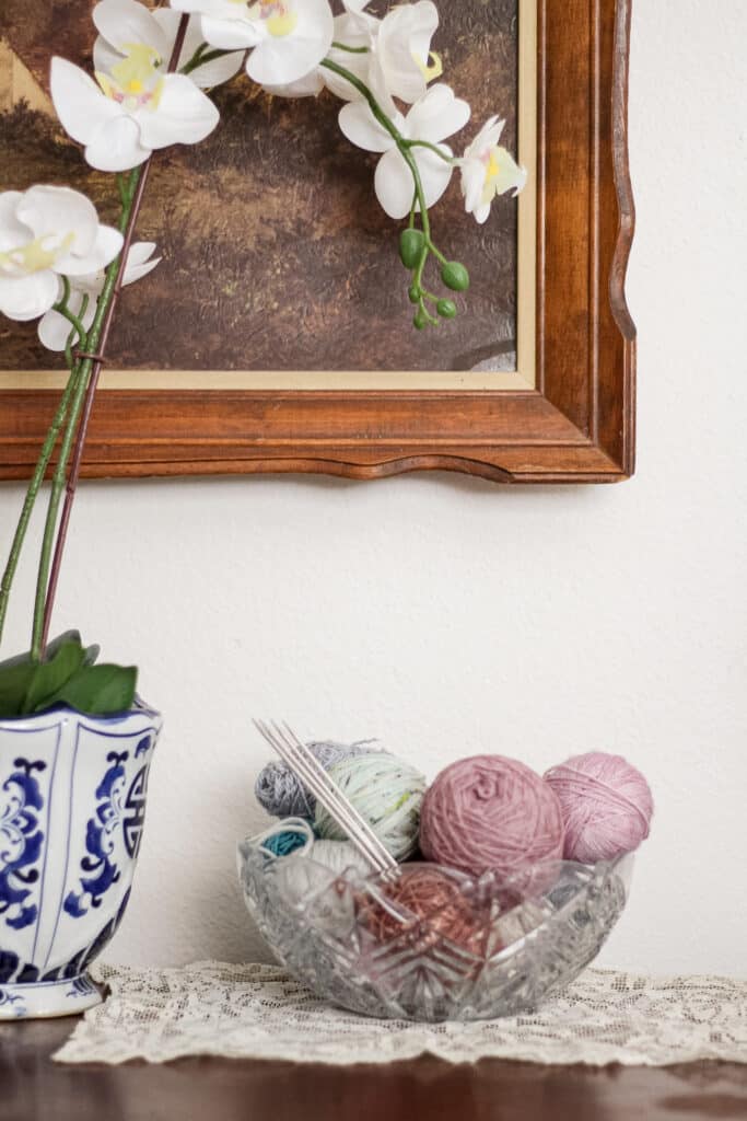 A crystal bowl filled with balls of leftover yarn and some metal double-pointed needles sits on top of a dark wood table next to a blue and white planter full of silk orchids.