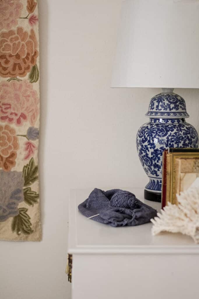 Some blue stockinette knitting sits on top of a white dresser. In the background are a blue and white lamp and a pastel tapestry hanging on the wall.