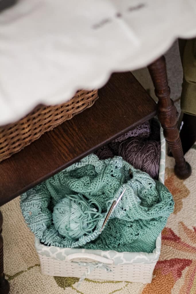 A pile of green and purple knitting in a white basket peeps out from under a dark wood table.