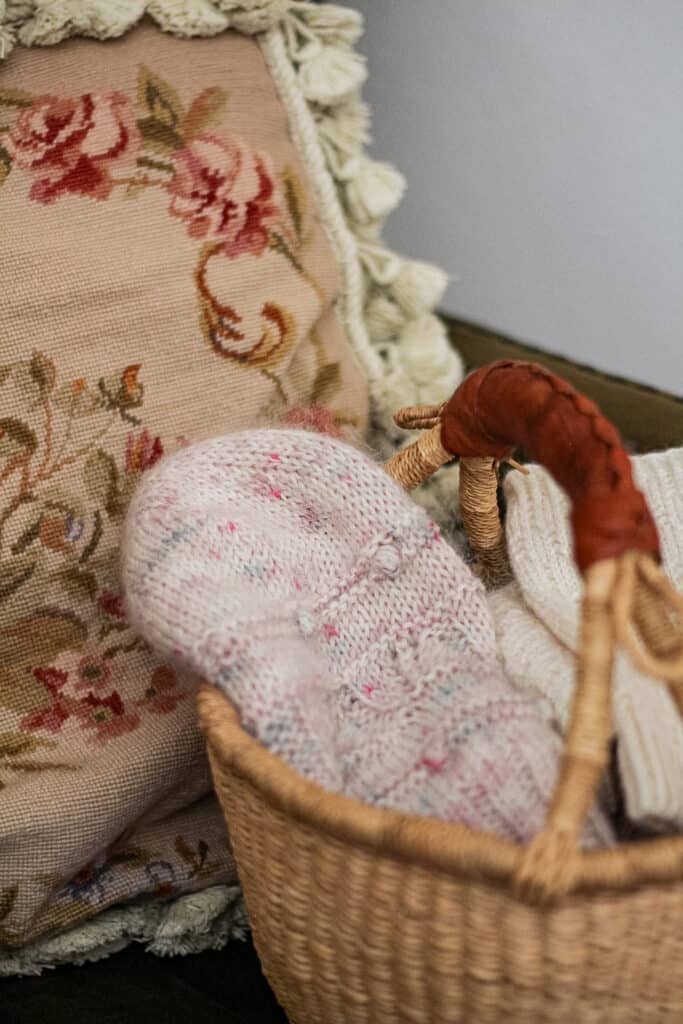 A pink, fuzzy, handknit hat with little bobbles sits in a woven basket on a green armchair in front of a needlepoint cushion.