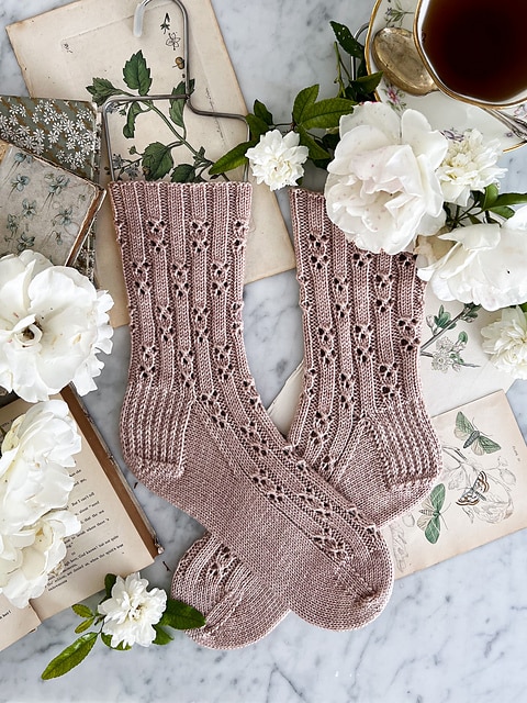 A pair of soft pink handknit socks is laid flat on a white marble countertop with the toes crossing each other. The socks have a repeating eyelet stitch pattern. They're surrounded by white roses and antique paper ephemera.